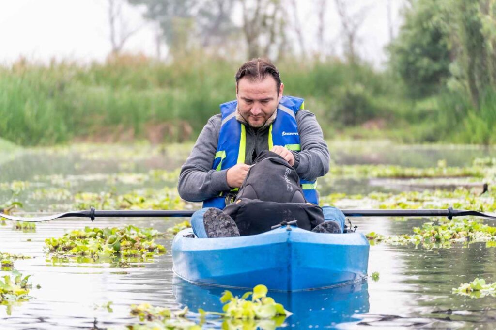 A man wearing a blue life jacket securing the roll top closure of a black dry bag while sitting in a kayak surrounded by lily pads