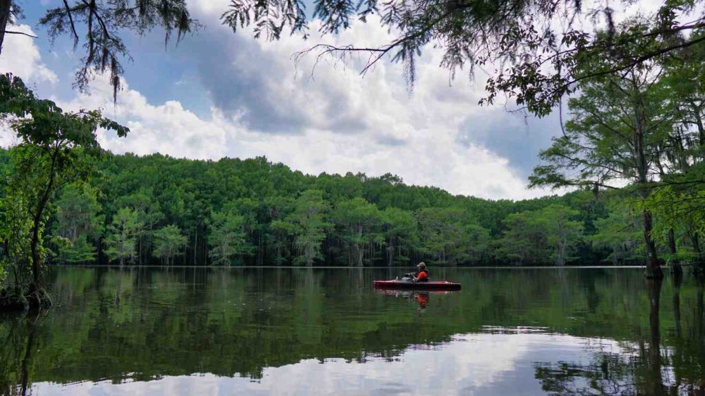 Person in red shirt fishing from a red kayak on a perfectly calm forest lake with green tree reflections