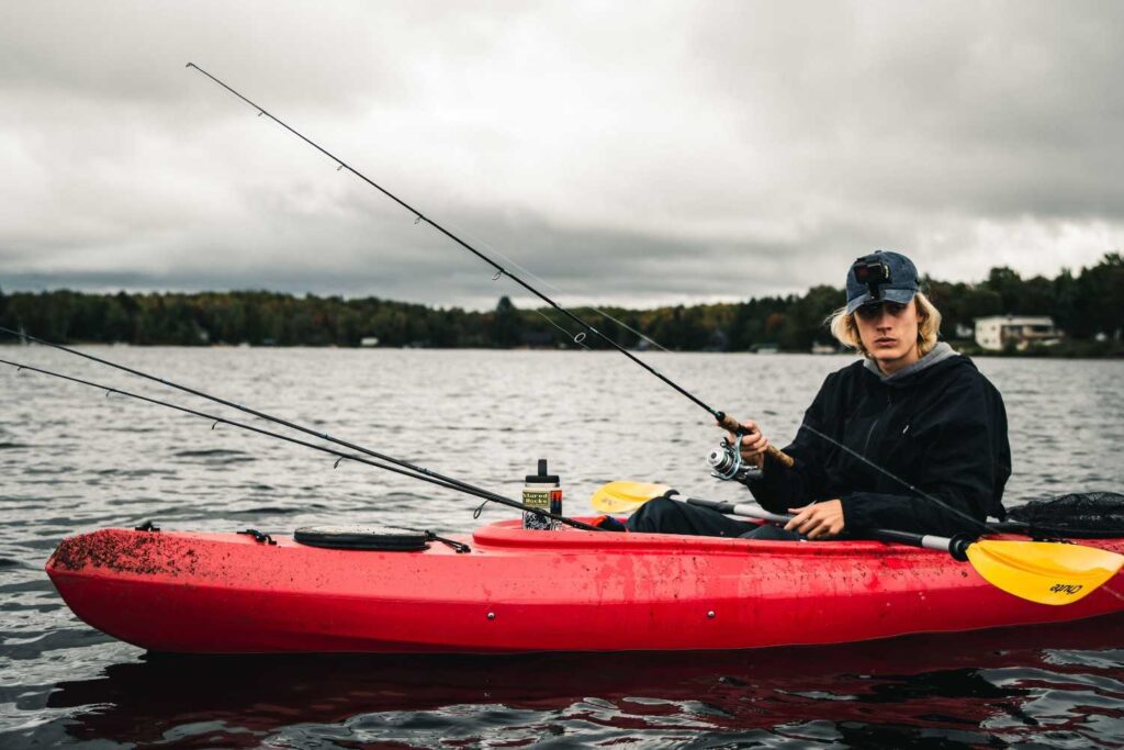 Young man holding fishing rod in a red kayak with multiple rods set up on a cloudy autumn lake