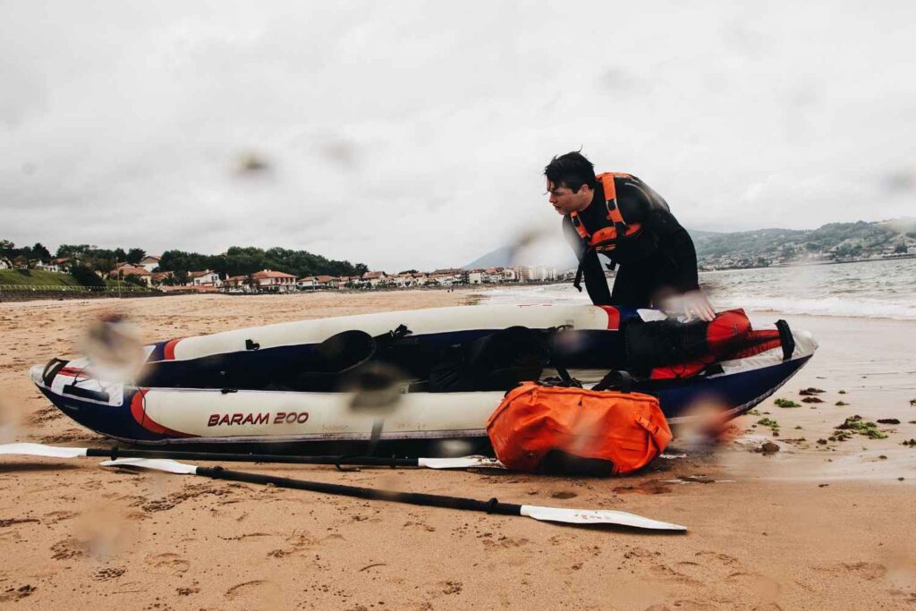 An orange dry bag resting on a sandy beach next to a Baram 200 inflatable kayak with a man preparing gear in the background