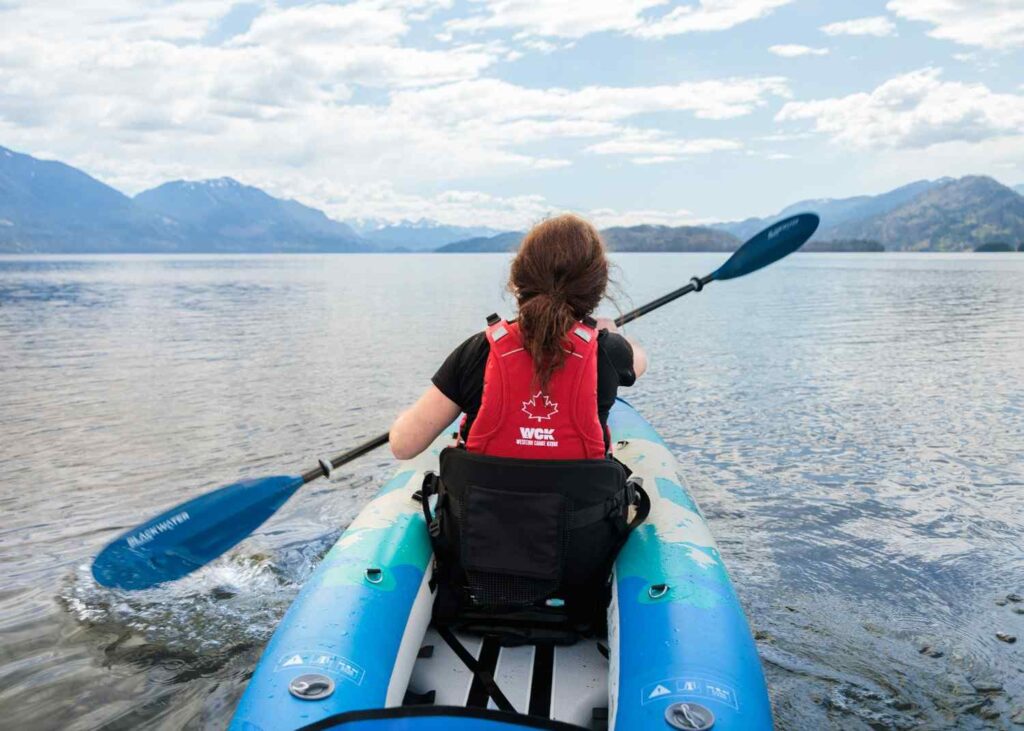A woman paddling a wide inflatable kayak with a high-back supportive seat on a calm mountain lake with snow-capped peaks in the background