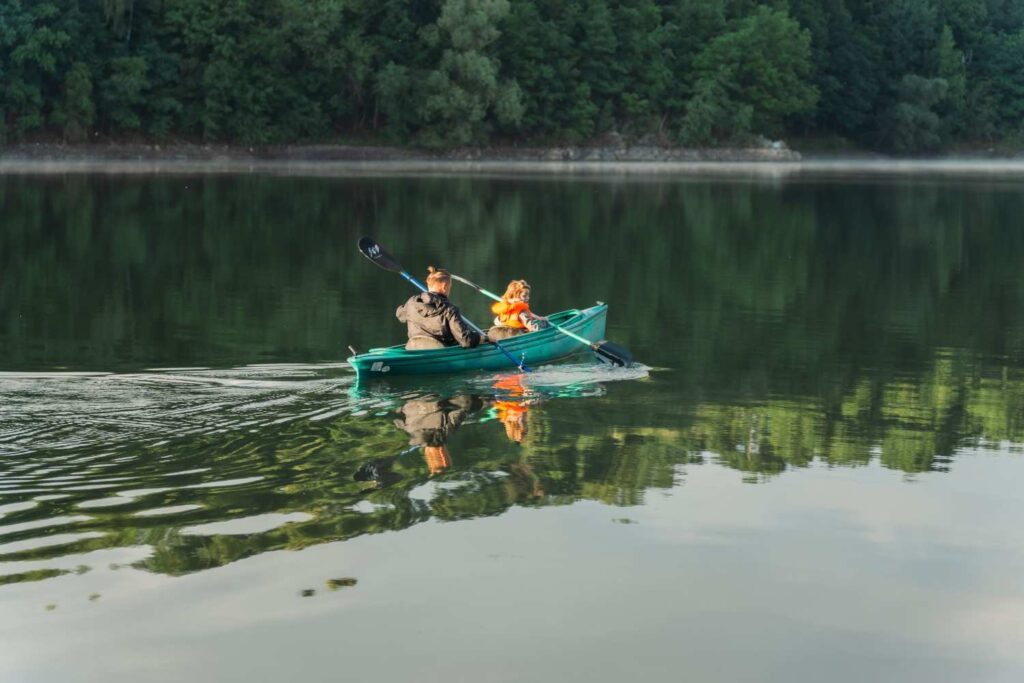 A parent and young child paddling together in a green kayak on a calm misty lake, child wearing an orange life jacket