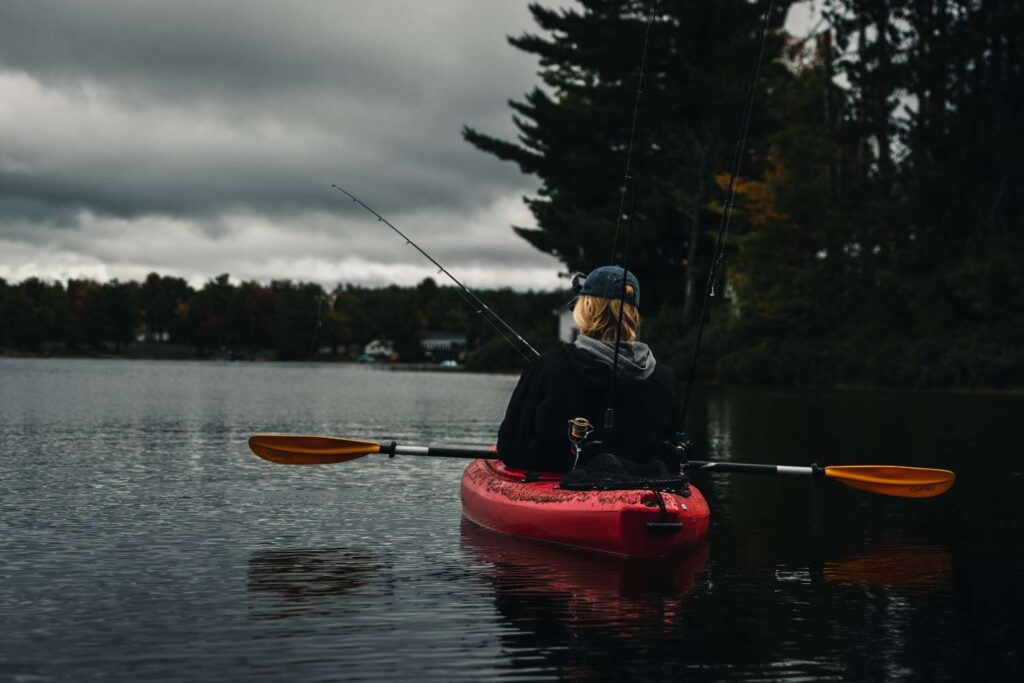Back view of a person sitting in a red fishing kayak with two rods deployed on a calm overcast lake