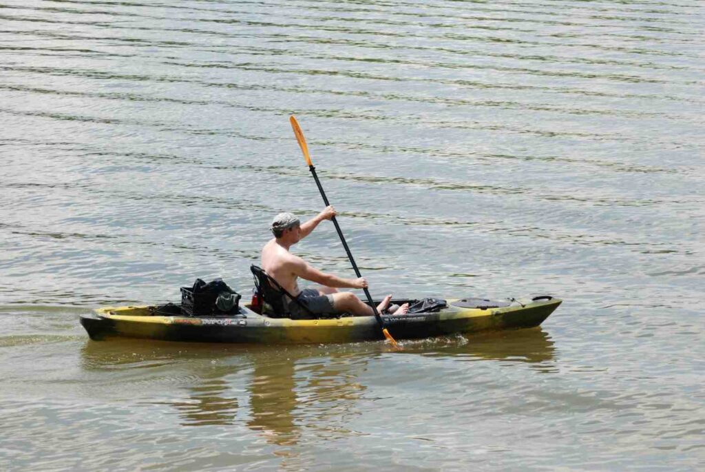 Shirtless man paddling a camo fishing kayak with gear loaded on a warm hazy summer day on calm water