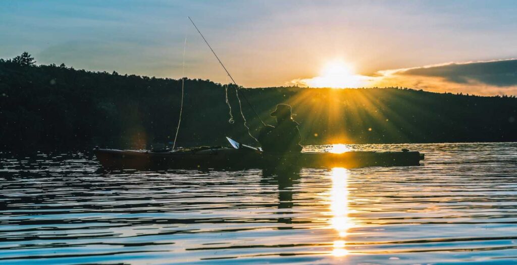Silhouette of a person fishing from a kayak with rod raised against a glowing sunset over the lake