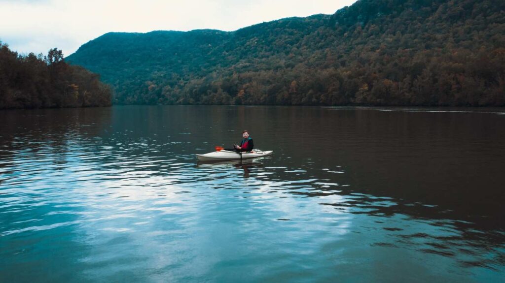 Person in a white kayak sitting alone on a calm dark mountain lake surrounded by autumn forested hills