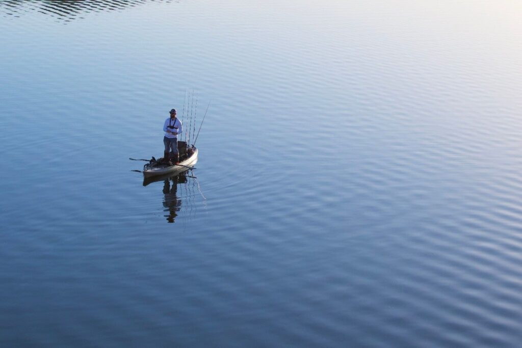 Man standing on a fishing kayak with several rods set up, casting on calm reflective water