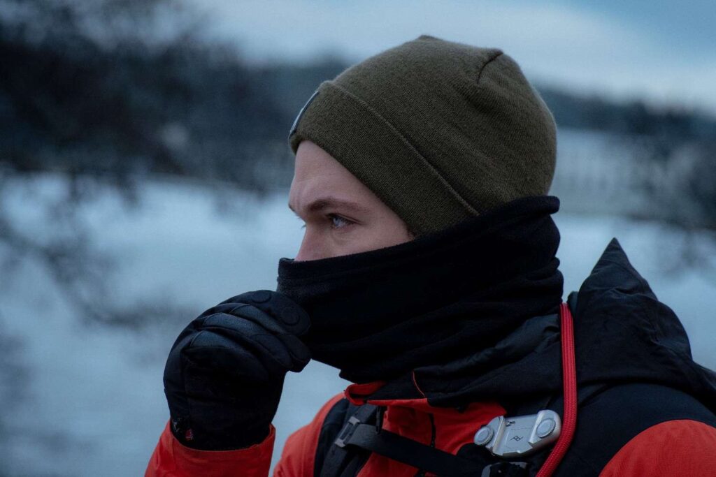 A close-up portrait of a man wearing a beanie, neck gaiter, gloves, and red jacket in a snowy outdoor winter setting