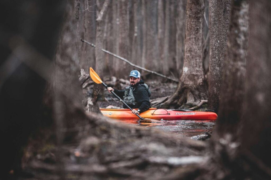 Man in fishing vest paddling an orange Wilderness Systems Pungo 120 kayak through a narrow dark forest creek