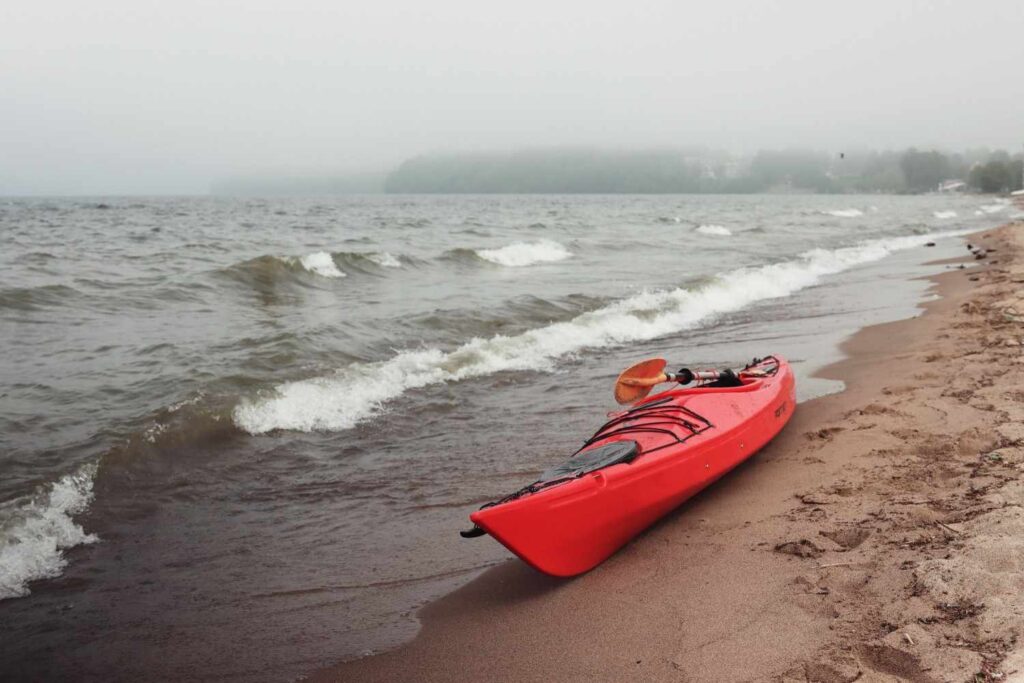 A red kayak resting on a sandy beach with rough choppy waves and thick fog in the background showing dangerous ocean conditions