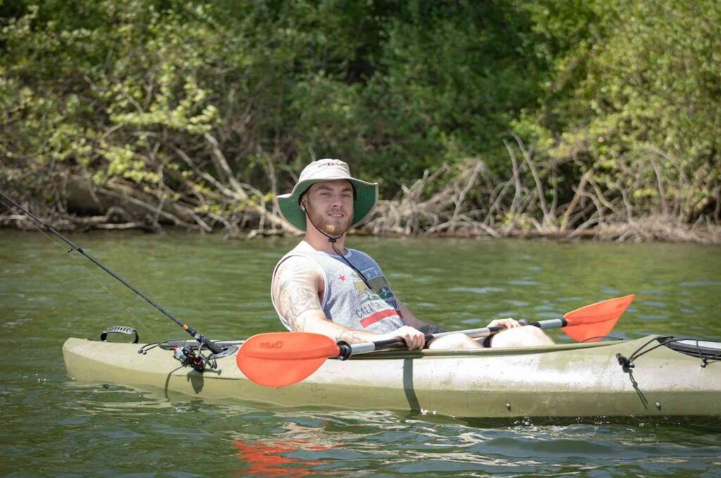 A smiling man in a fishing kayak with one rod near green shoreline vegetation on a calm freshwater river
