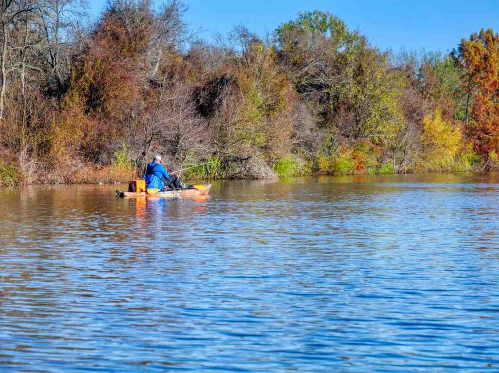 A man fishing from an orange pedal fishing kayak on a calm lake surrounded by colorful autumn trees with a gear crate and fishing rod mounted