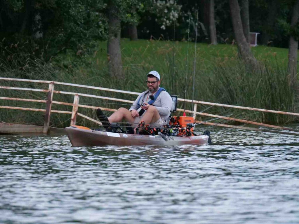 A man sitting in a heavily rigged tan pedal fishing kayak with multiple rods, orange gear crate, fish finder, and full tournament setup on a lake