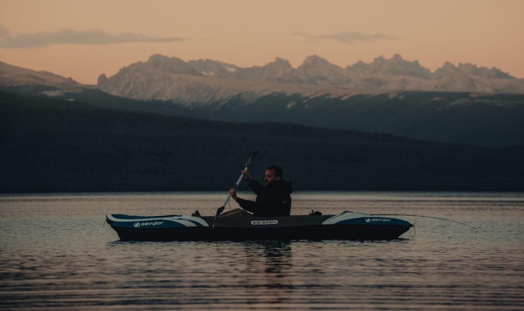 A man paddling a Sevylor Big Basin inflatable kayak on a calm mountain lake at dusk with snow-capped peaks in the background