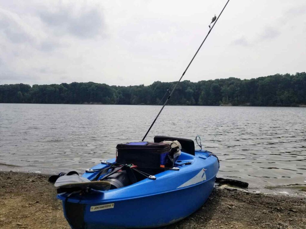 A blue fishing kayak resting on a muddy lake shore with a single fishing rod, tackle bag, and basic gear setup on a cloudy day
