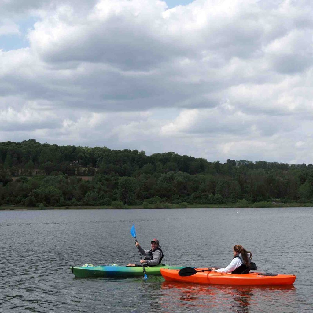 Man and woman paddling separate sit-on-top kayaks on a calm lake under cloudy sky with forested hills behind