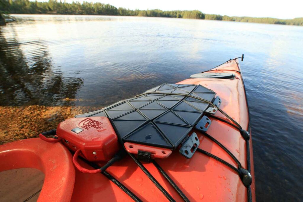 A close-up of a solar charging panel and waterproof battery box strapped to the deck of an orange kayak on a calm lake
