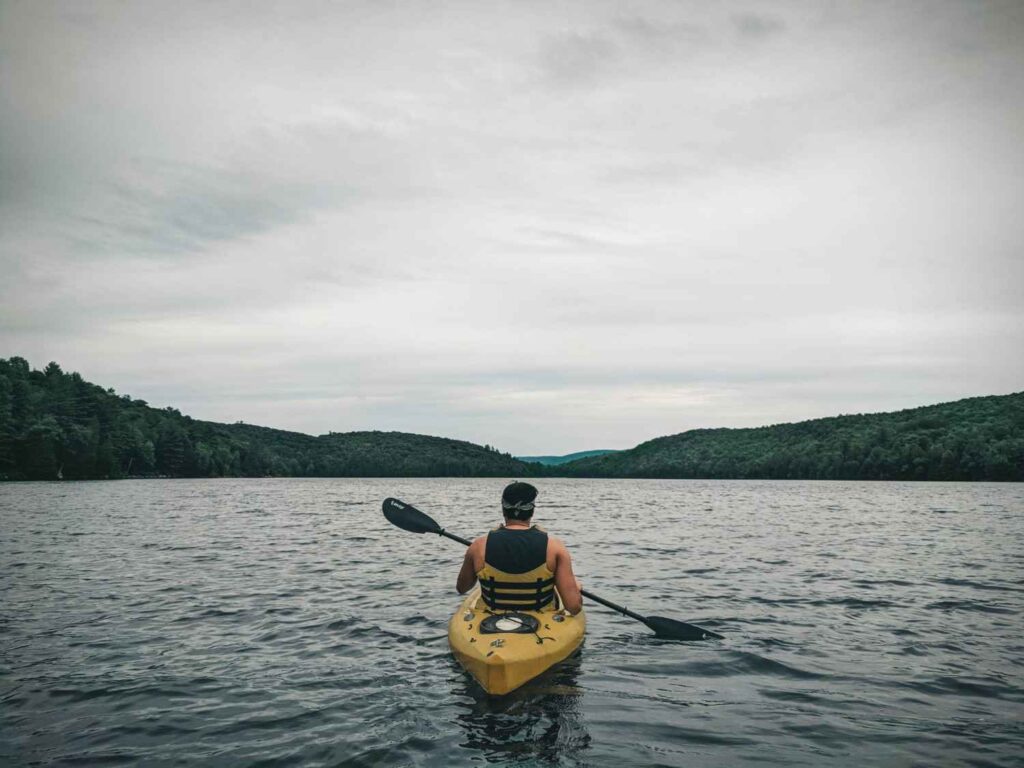 A man paddling a yellow kayak away from the camera on a calm grey lake surrounded by forested hills under overcast skies