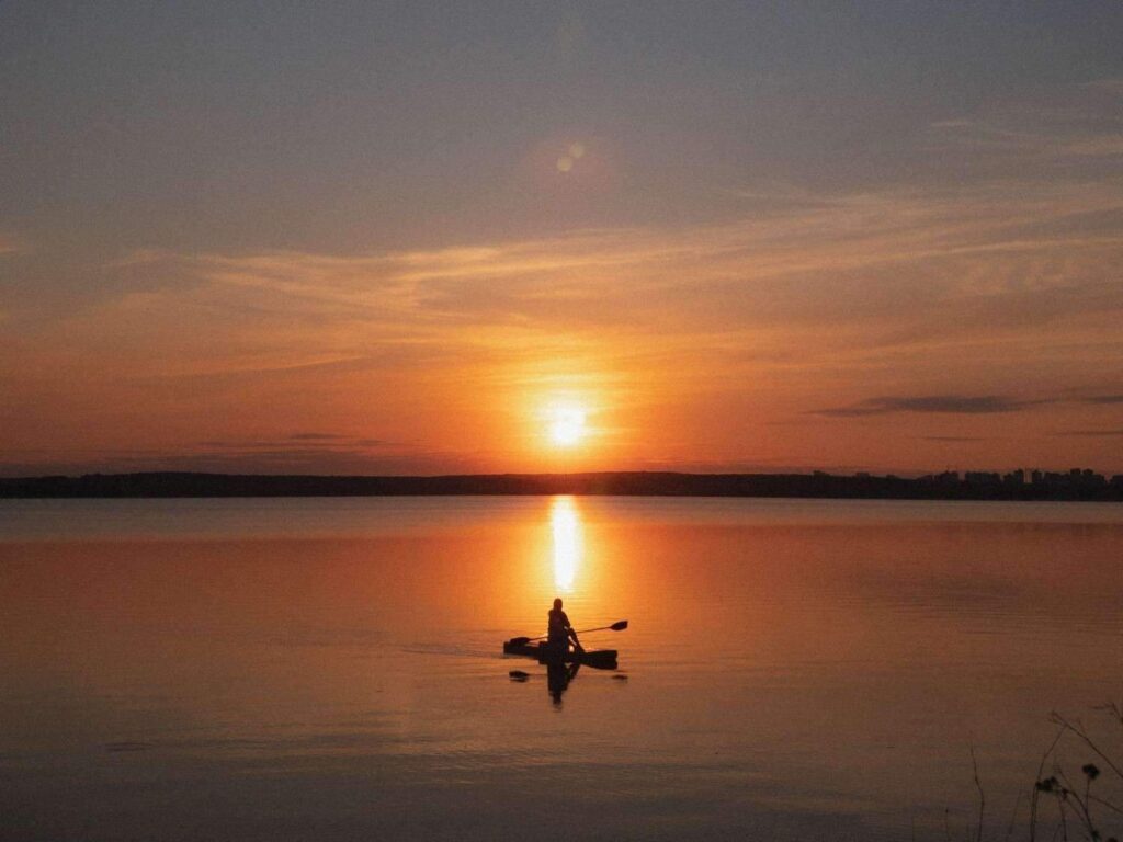 Silhouette of a solo kayaker paddling on perfectly calm water with a dramatic golden sunset reflected on the surface