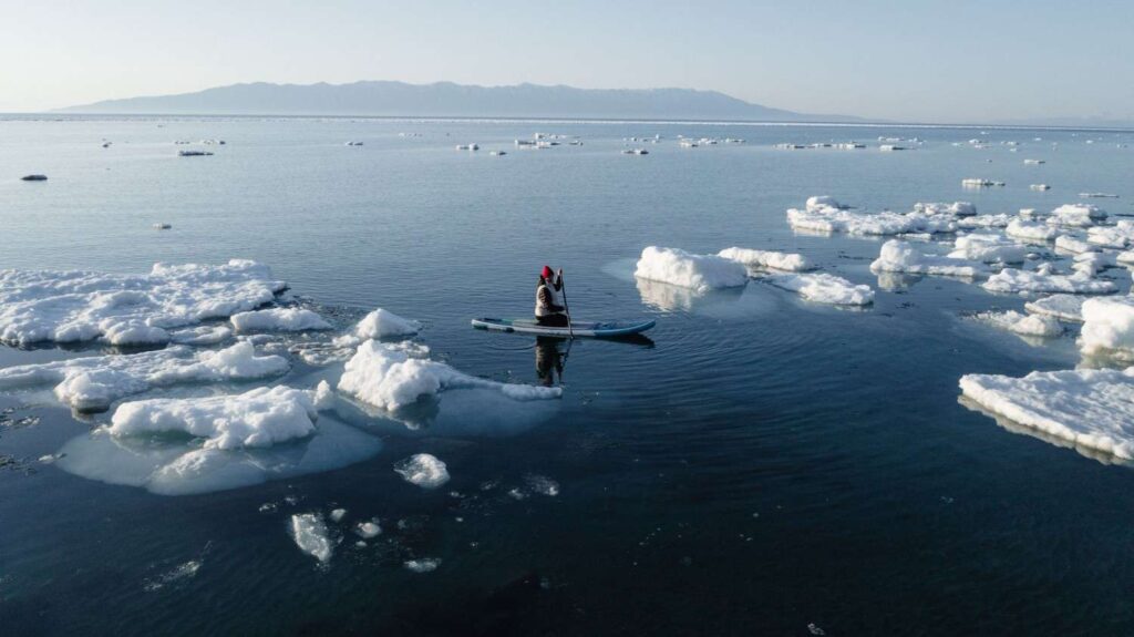 A lone paddler in winter gear navigating calm water surrounded by floating ice chunks with mountains in the distance