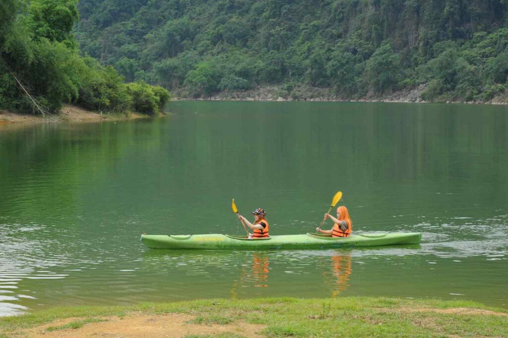Two people wearing orange life jackets paddling a green tandem kayak together on a calm green lake surrounded by lush forest