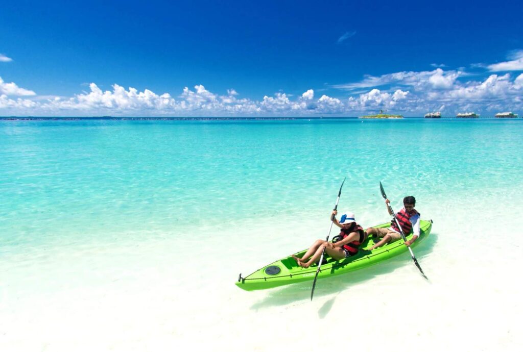 Man and woman in life jackets paddling a green tandem kayak over clear turquoise water near a tropical island