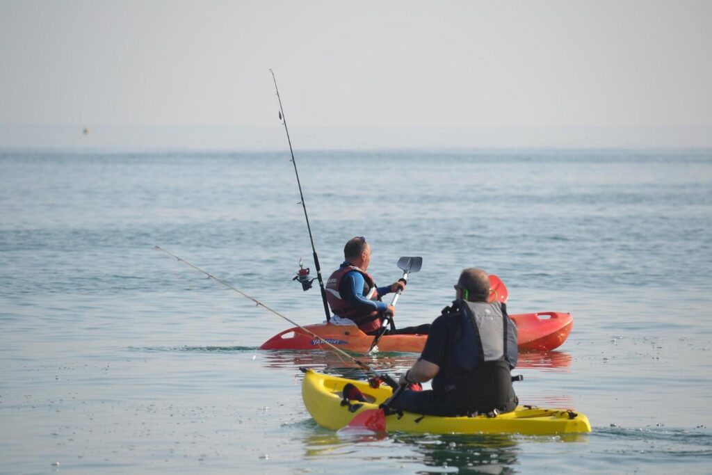 Two anglers paddling wide sit-on-top fishing kayaks on calm open water with fishing rods mounted and life vests on