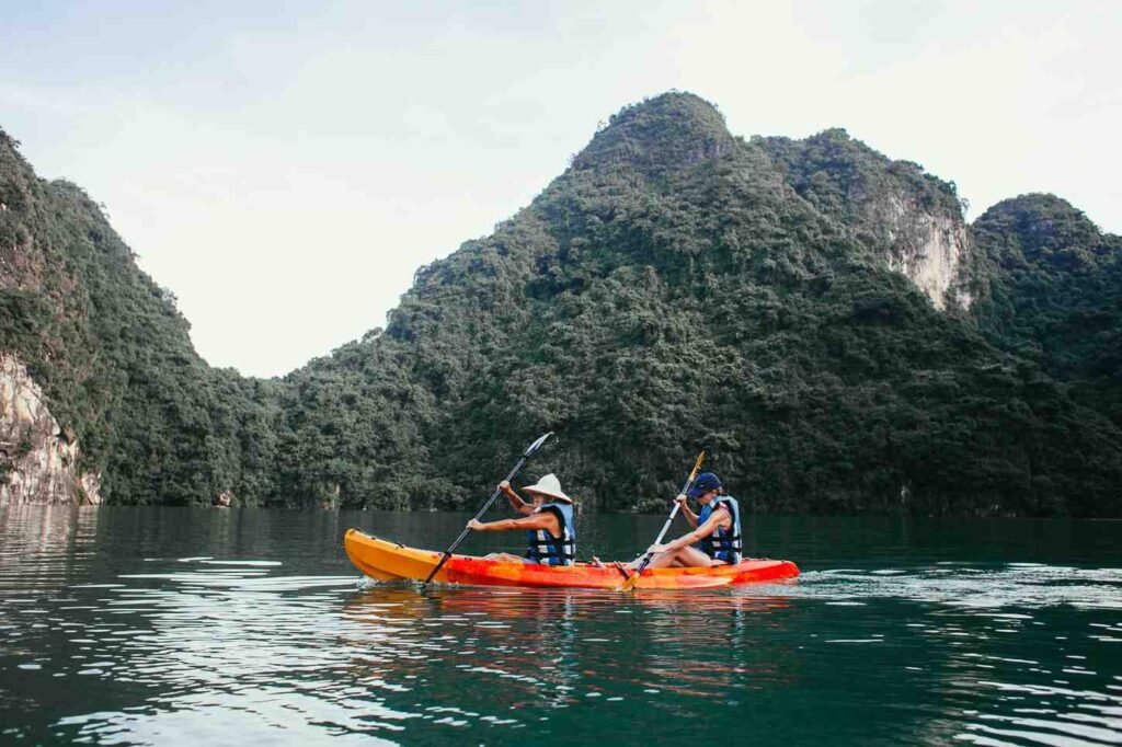 Two kayakers paddling sit-on-top kayaks on calm water with lush green mountains in the background