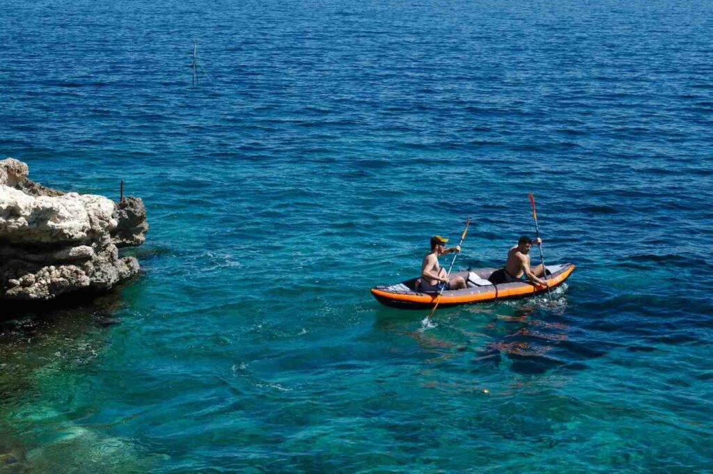 Two men paddling an inflatable tandem kayak on clear turquoise water beside jagged white limestone rocks on a sunny day