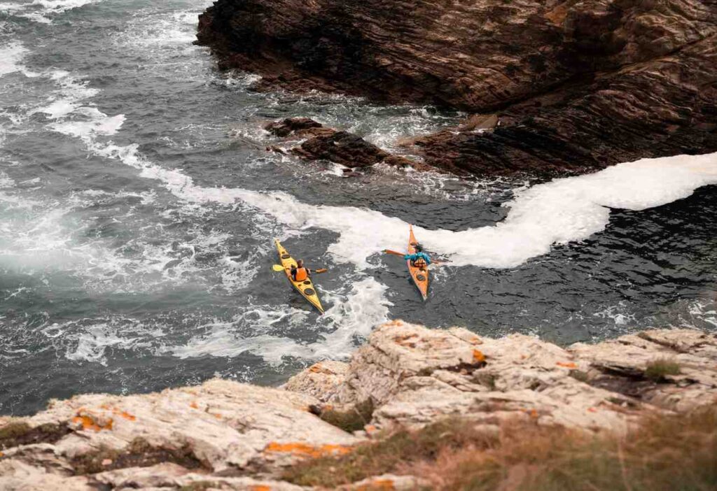 Aerial view of two sea kayakers paddling through churning white water near jagged coastal rocks showing how to read dangerous ocean conditions