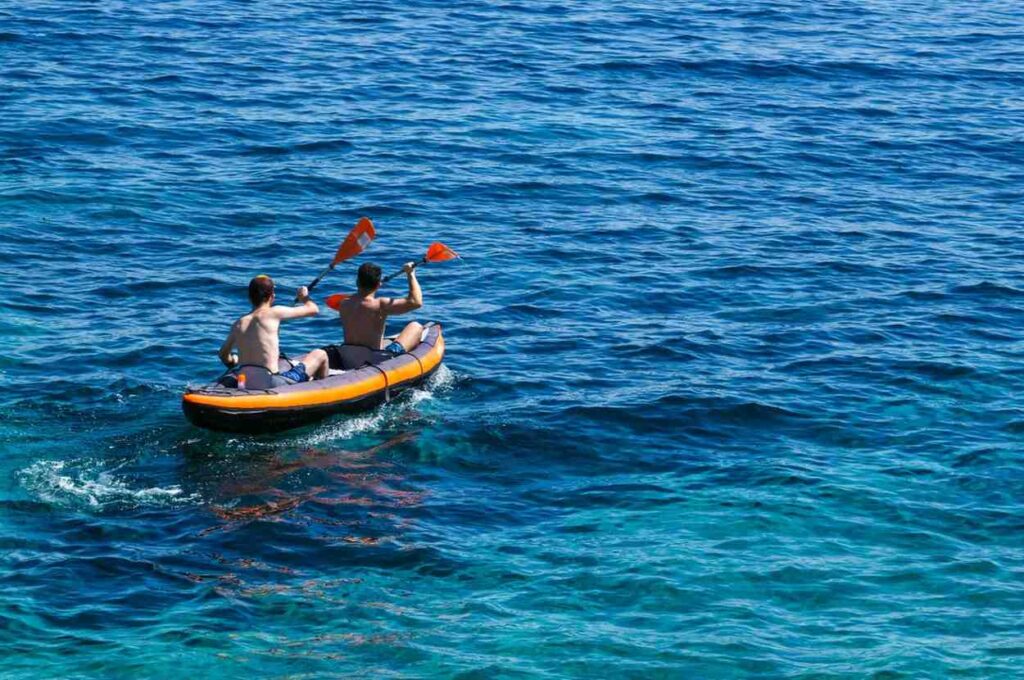 Two men paddling an orange and black inflatable kayak across choppy open ocean water with splashing waves