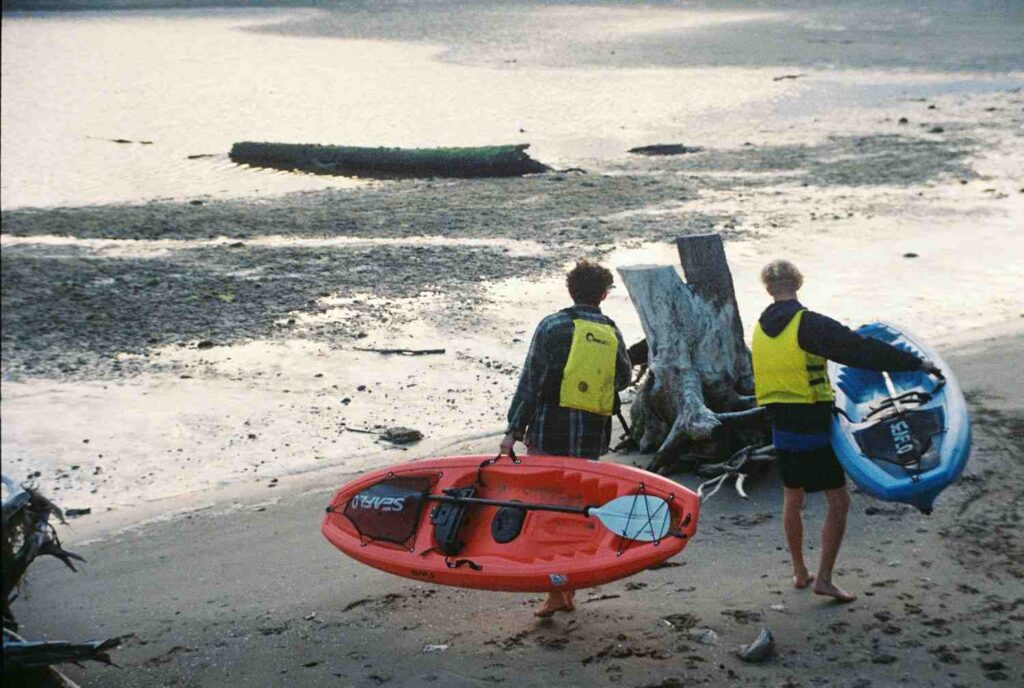 Two people in life vests carrying kayaks across a sandy beach toward the water at dusk, showing the physical effort of kayak transport