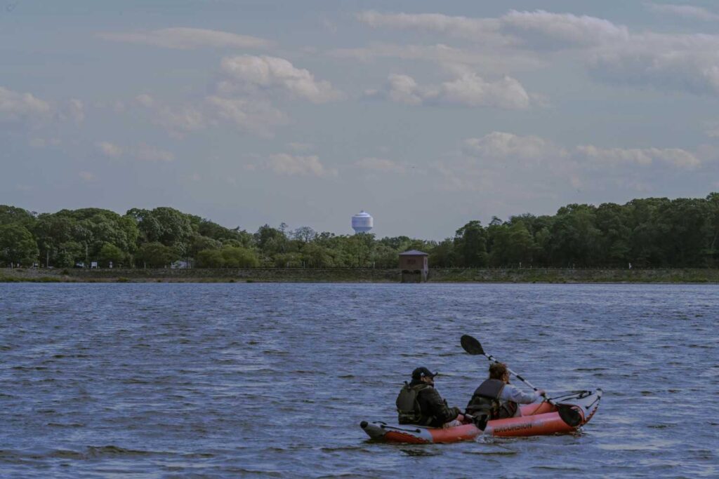 Two people paddling an inflatable Intex Excursion kayak on a windy open reservoir with choppy water and a water tower visible in the background