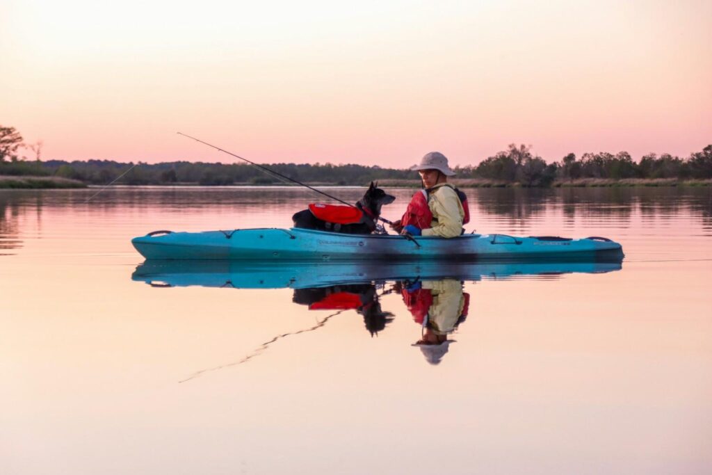 A woman with her dog on a blue fishing kayak on glassy calm water at sunset with a fishing rod, surrounded by beautiful pink sky reflection