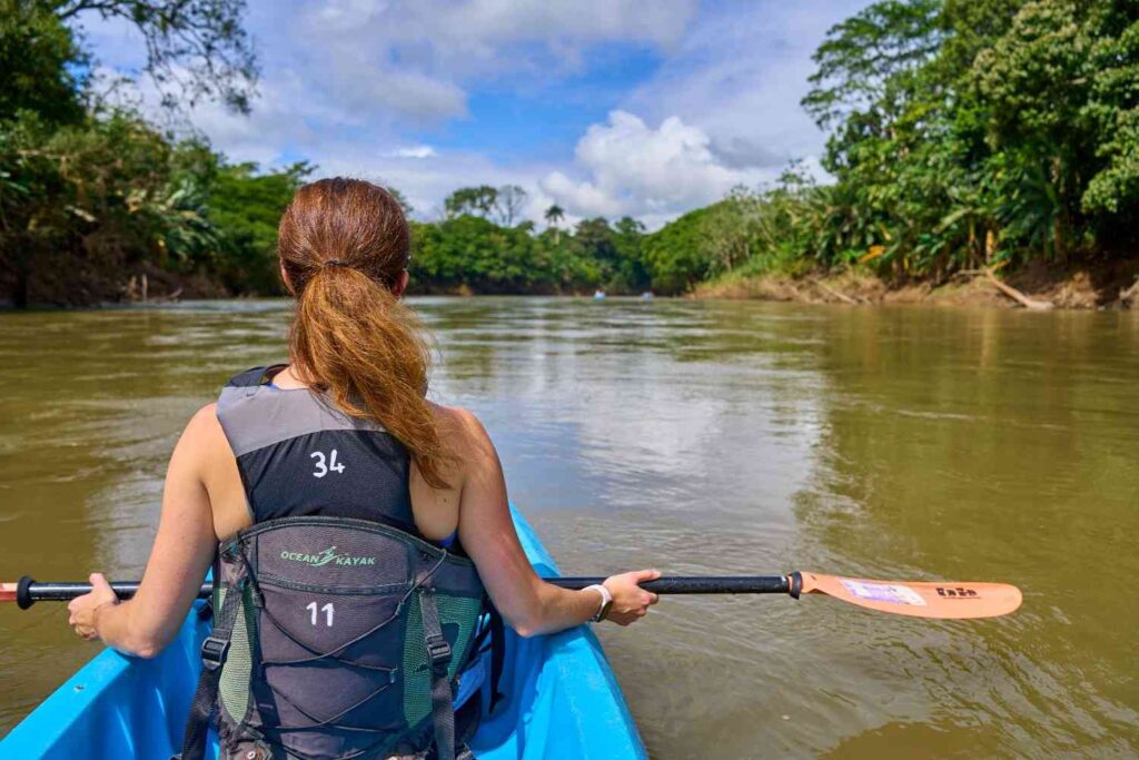 A woman wearing a high back Ocean Kayak PFD paddling through a tropical river with lush jungle vegetation on both sides