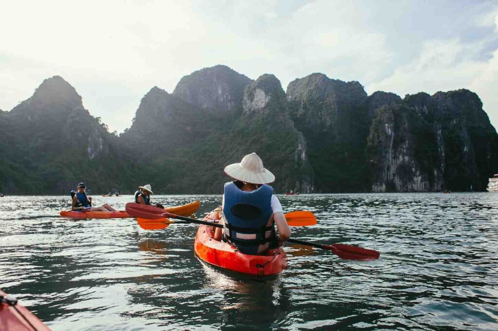 A woman wearing a blue life jacket paddling a kayak through a scenic bay surrounded by dramatic limestone mountains with other kayakers in the background