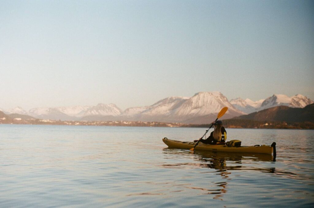 A woman paddling a yellow kayak on a glassy lake with snow-capped mountains in the background at golden hour