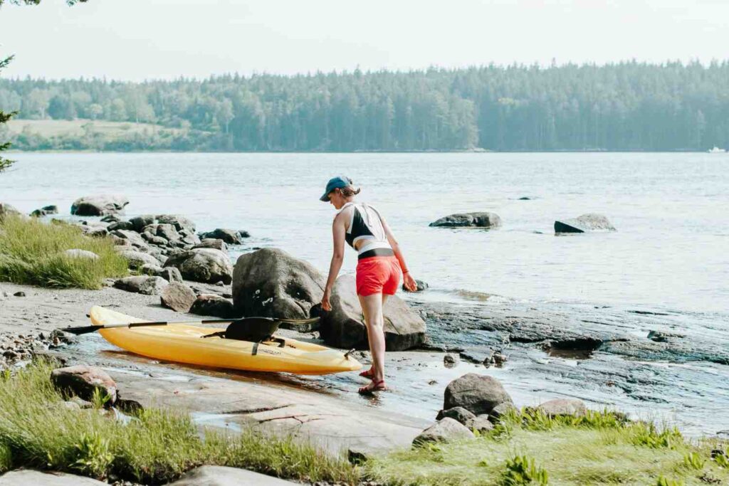 A woman solo launching a yellow kayak on a rocky shoreline near calm water, demonstrating easy single-handed kayak transport