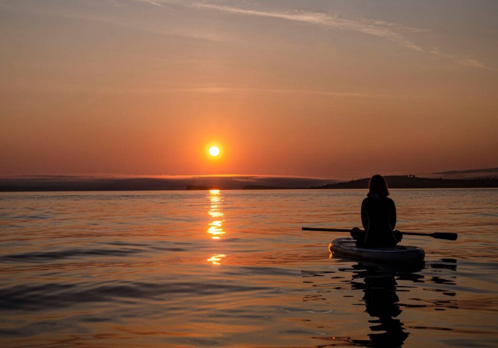 A woman sitting on a paddleboard holding a paddle watching a stunning orange sunset reflected on a calm open lake