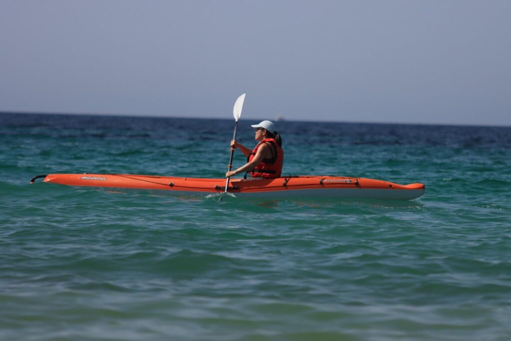 A woman in an orange sea kayak with life jacket paddling alone on open turquoise water far from shore