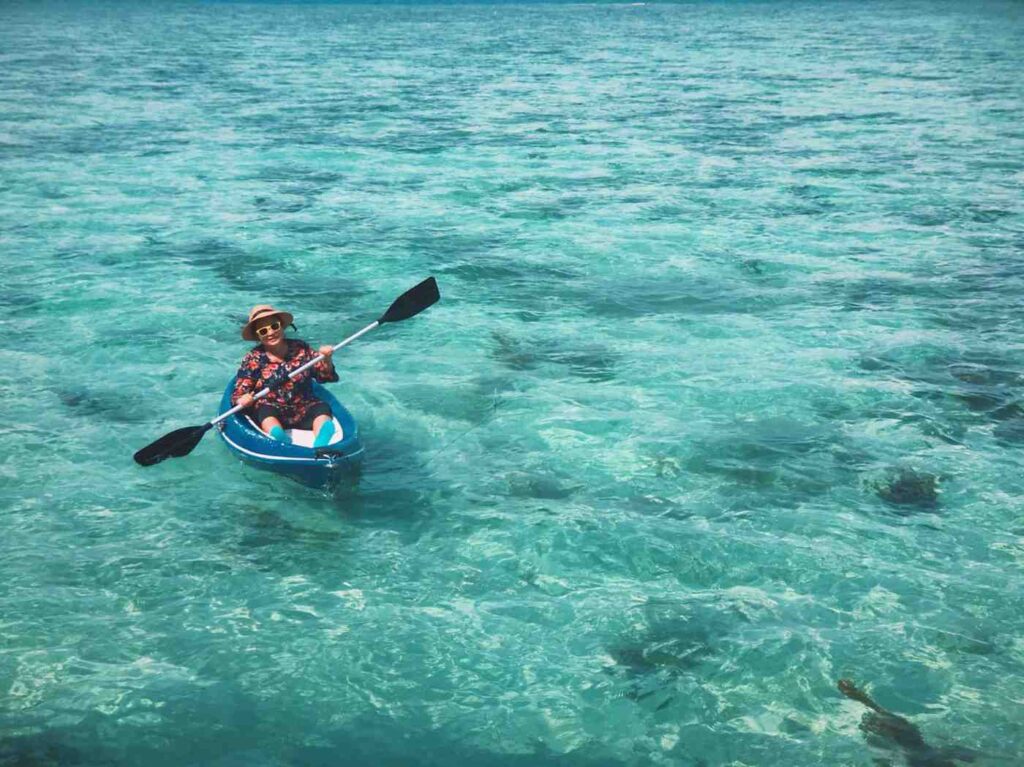 A woman wearing a sun hat paddling a blue sit-on-top kayak across crystal clear turquoise ocean water