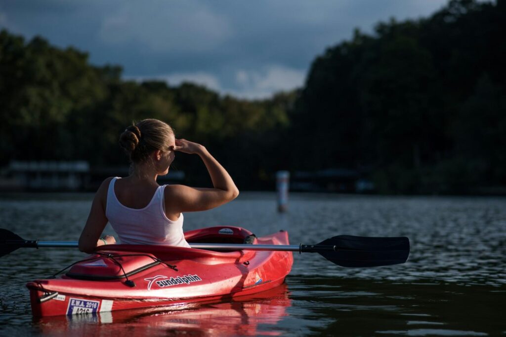 A young woman sitting in a red Sun Dolphin kayak on a dark lake shielding her eyes while looking into the distance