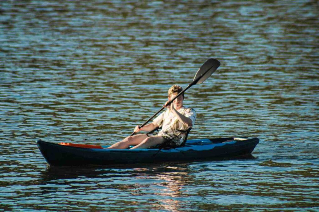 A young man paddling a sit-inside kayak alone on calm open water in casual clothes on a sunny day