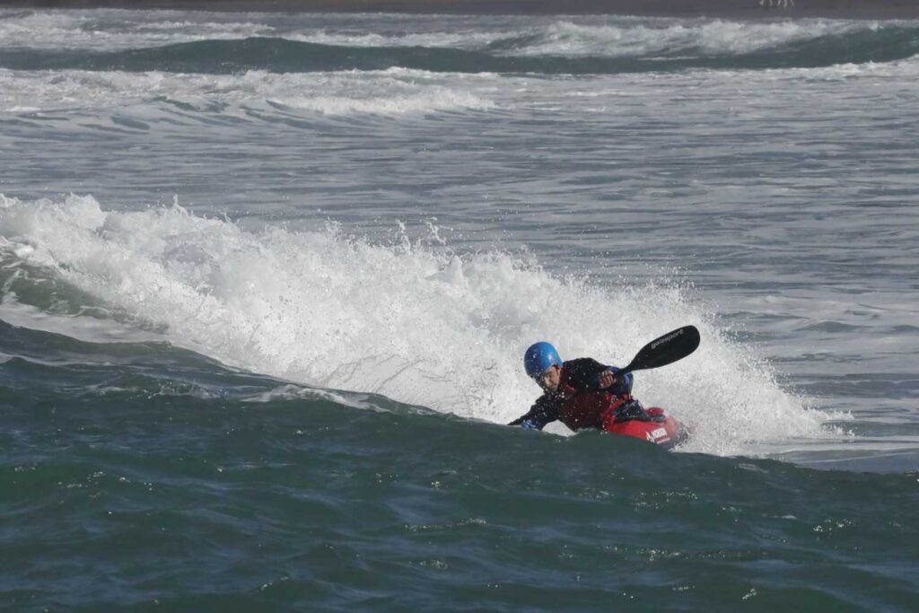Kayaker paddling through a large breaking wave, helmet and gear visible amid heavy splash
