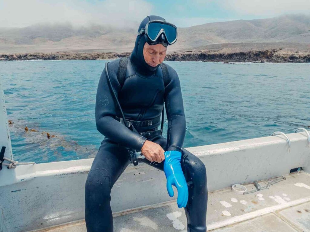 Person in a wetsuit pulling on a blue neoprene-style glove while sitting on a boat