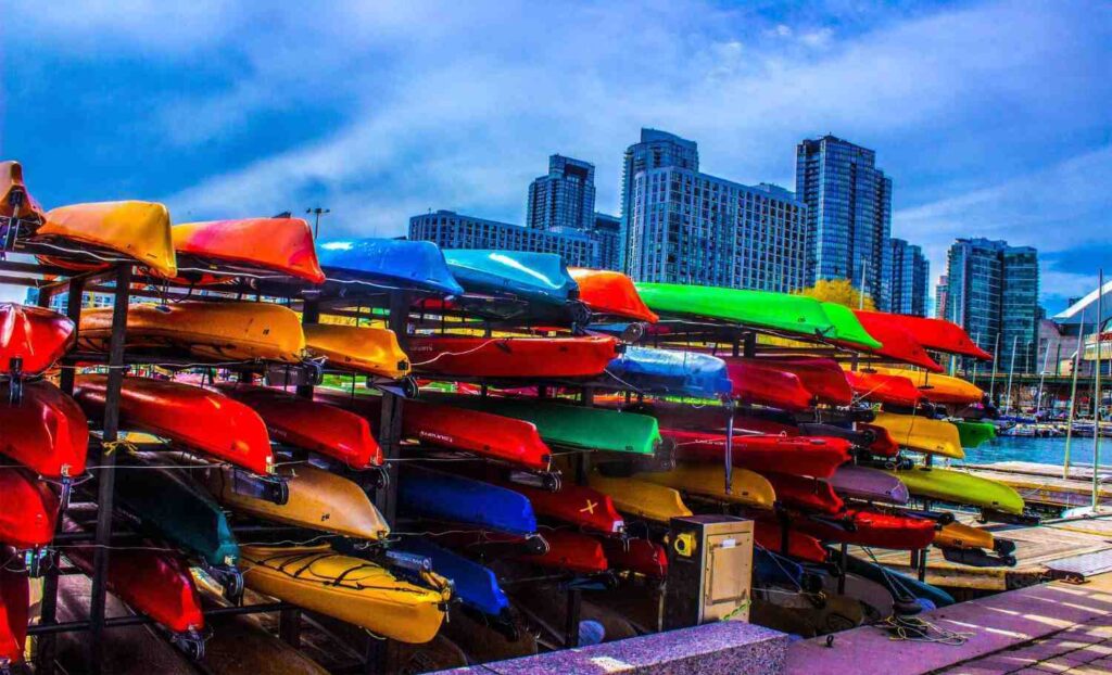 Multiple colorful kayaks of different types stacked on racks with a city skyline in the background