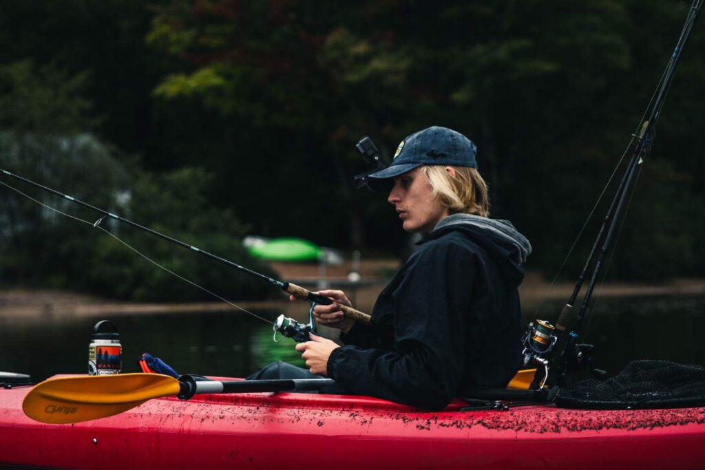 Person fishing from a red sit-on-top kayak with multiple rod holders, net, and gear within easy reach