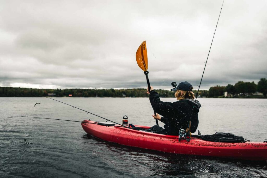 An angler in a red fishing kayak lifting the paddle mid-stroke with rod holders and tackle visible