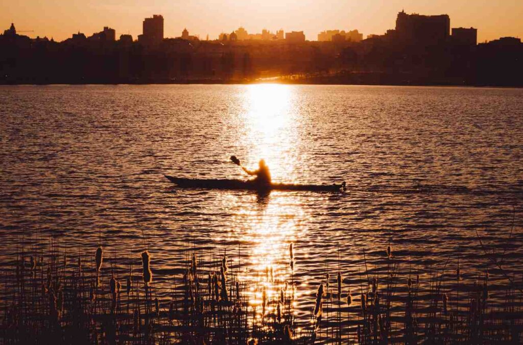Silhouette of a kayaker paddling across calm water at sunset with a city skyline in the background