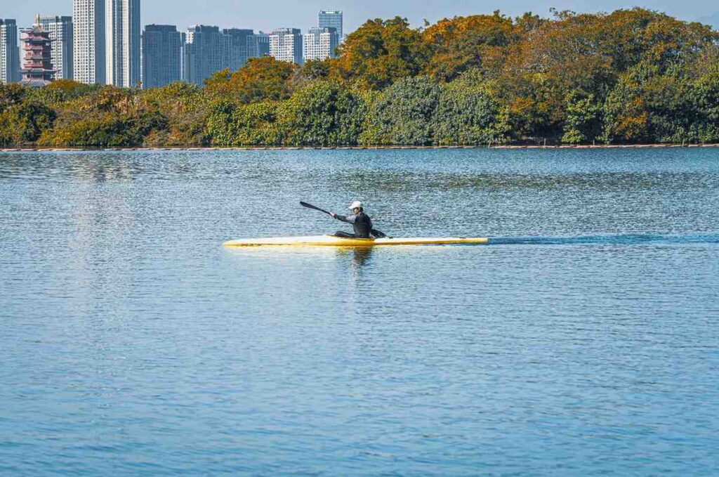 Man paddling a sleek narrow kayak in a straight line across calm open water with city skyline in background